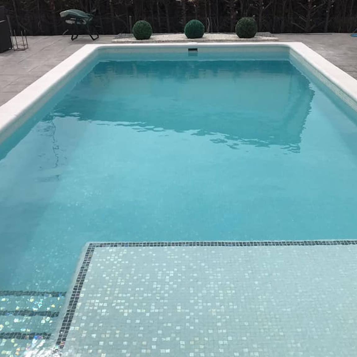 Rectangular pool with mosaic tiles on the edge, surrounded by a wooden deck and plants.